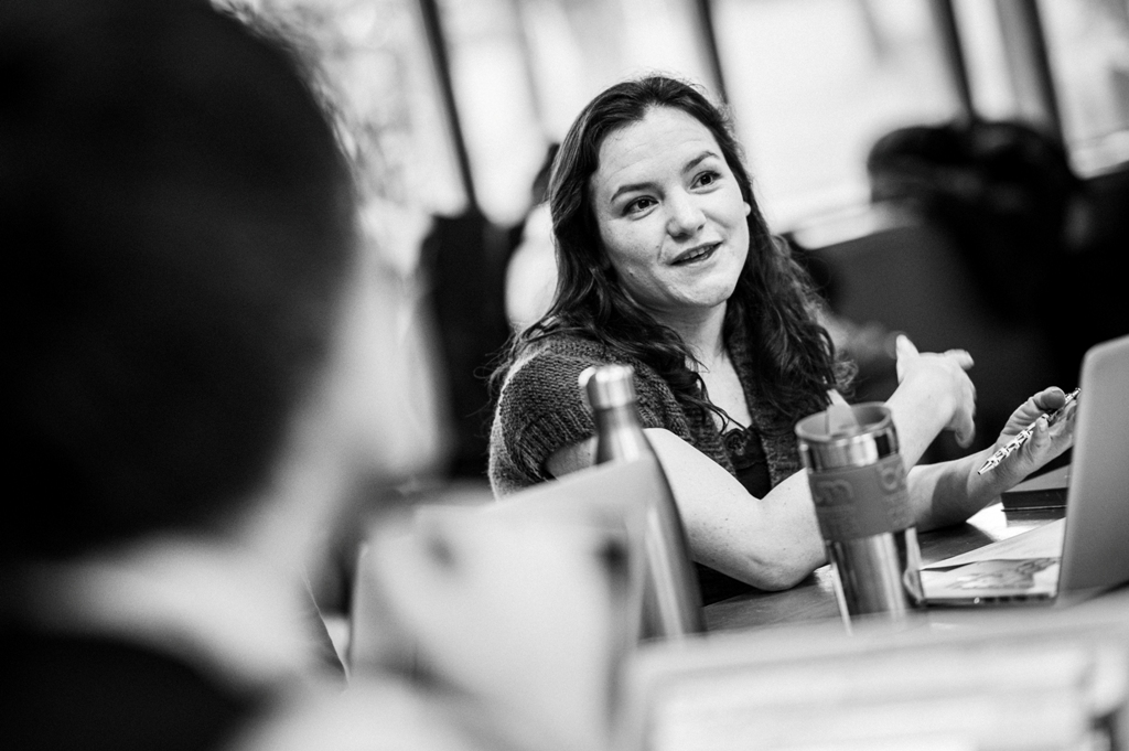 Black-and-white portrait of Laura Jones in conversation with other people sitting at a table. The others are blurred or out of shot so that the focus is solely on Laura.