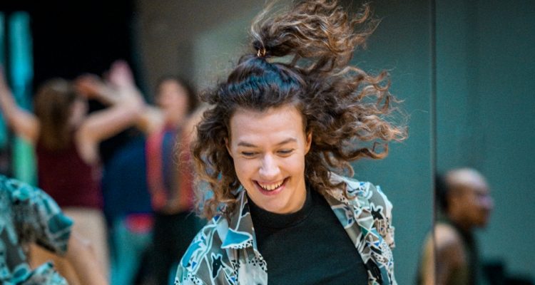 Portrait of Millie dancing in the studio, her long, brown, curly hair flying mid-movement. Millie is looking down, laughing.