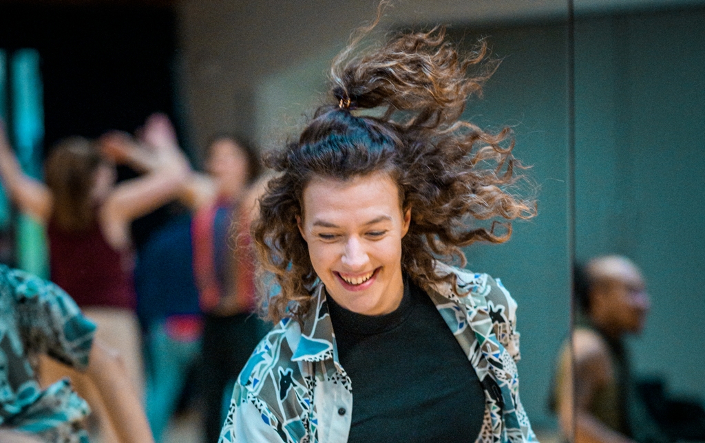 Portrait of Millie dancing in the studio, her long, brown, curly hair flying mid-movement. Millie is looking down, laughing. Photo by Chris Parkes.