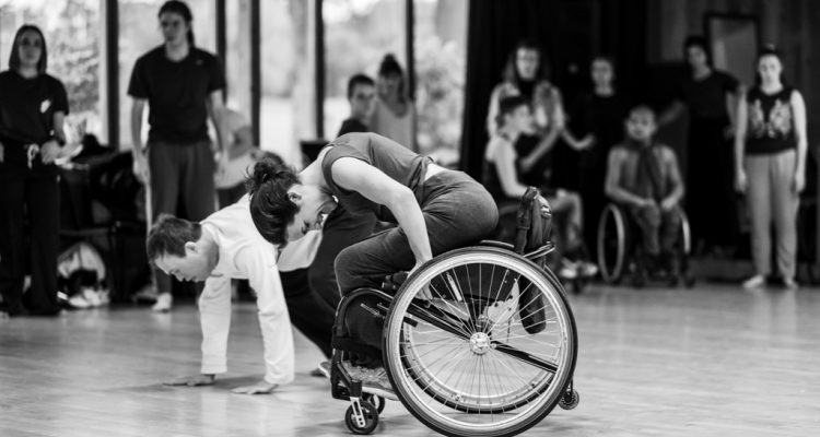 Black-and-white photo of Laura Jones dancing in her wheelchair in the studio. She is diving forward, gripping the wheels of her wheelchair. Chris Pavia is on all fours on the floor behind her, and in the background other dancers are watching.