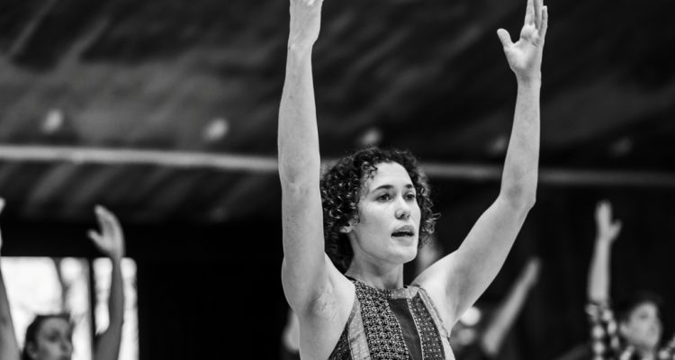 A black and white photo of Amy Butler leading class in a studio, she has both arms raised.