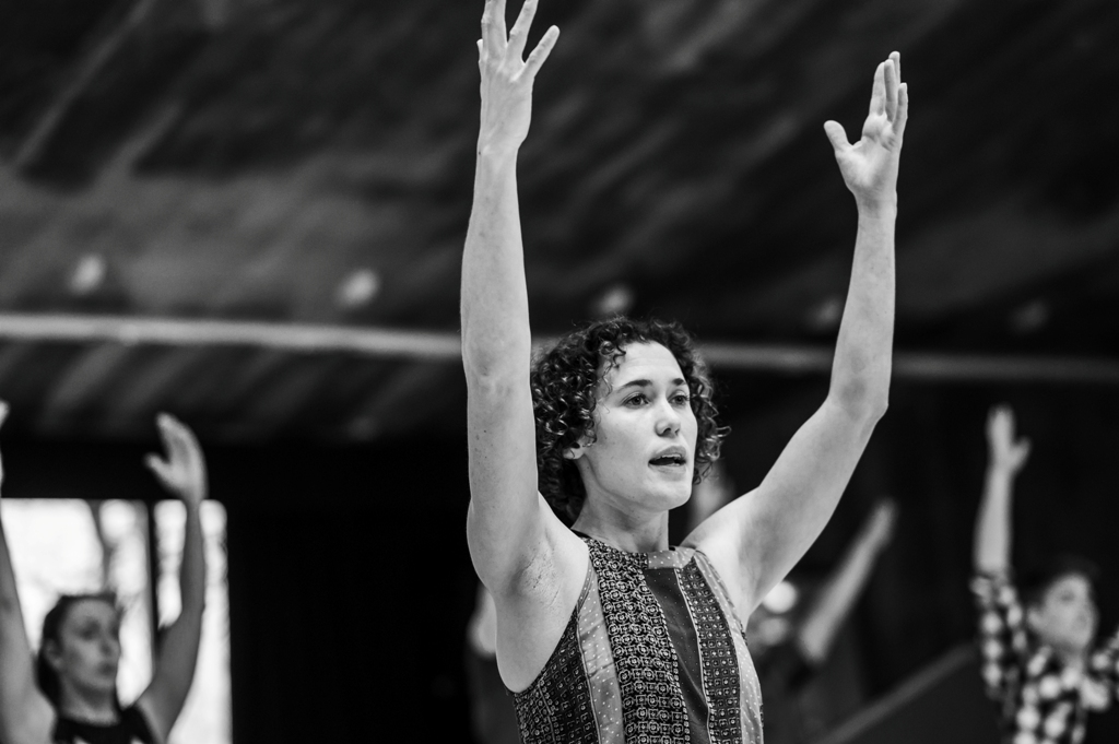 A black and white photo of Amy Butler leading class in a studio, she has both arms raised.