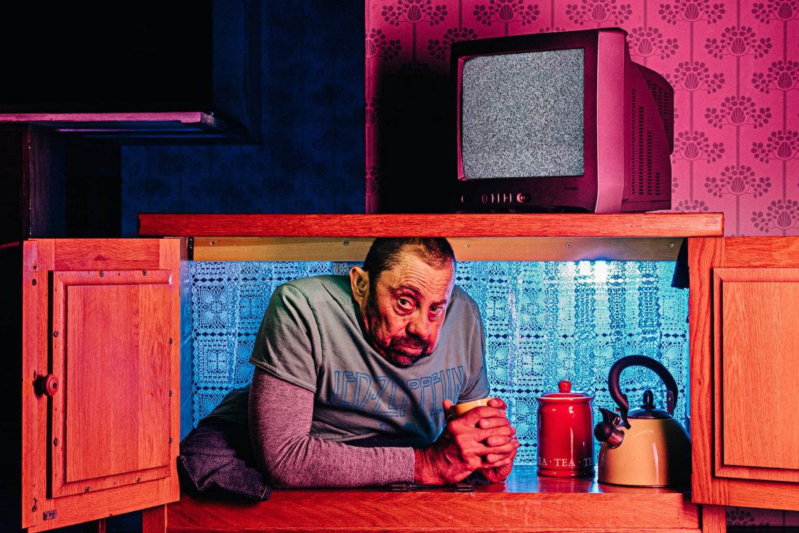 Dave Toole sits in a cupboard, pink and blue lights tint the image.