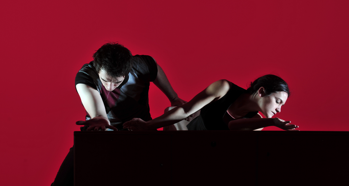 Two dancers at a table, one leans forward the other leans outward. With a deep red background.