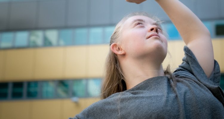 Hannah dancing outside in front of a tall building with large windows. She lifts one arm overhead and looks up towards her hand.