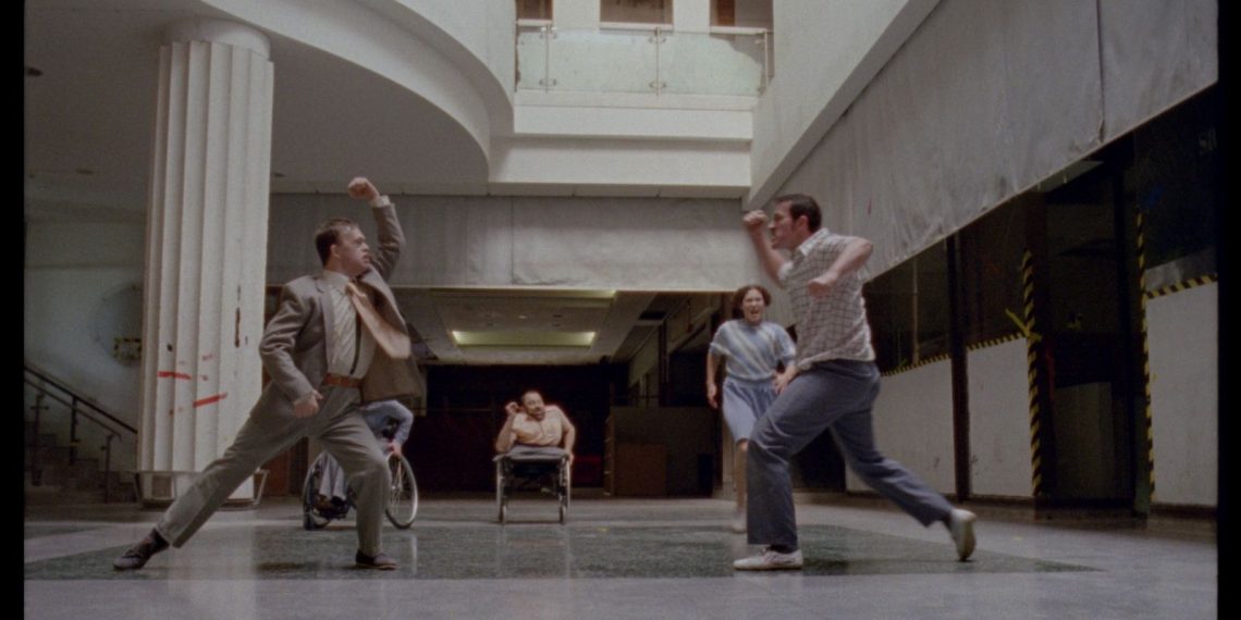 Film Still from Artificial Things. The cavernous inside space of a derelict shopping mall. On the left, a white column with vertical grooves supports a mezzanine level. In the centre, dancers Chris Pavia (left) and David Willdridge (right) face each other aggressively, fists raised. Amy Butler is running towards David from the back. In the background, Laura Jones and Dave Toole observe the scene, sitting in their wheelchairs.
