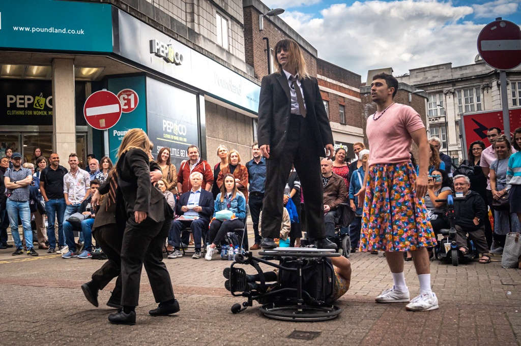 Outdoor performance of Frock, In the centre, Nadenh is lying on his side in his wheelchair. KJ is standing on the top wheel, wearing a black suit, white shirt and tie. To their left, two female dancers in black suits and ties are twisting around each other. To their left, Christian stands and looks at KJ, wearing a pink t-shirt and floral skirt.