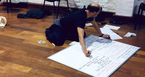 A photo of Tom Goodwin, kneeling on a wooden floor writing on a big whiteboard.