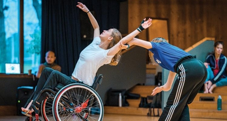 A wheelchair and a standing dancer dueting in the studio.
