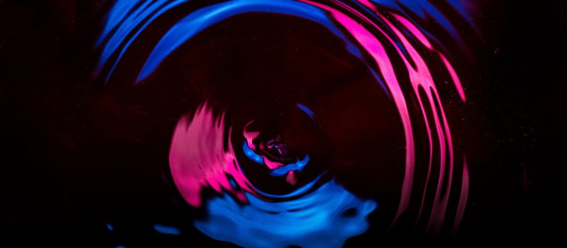 Looking down from above, concentric circles ripple out from a water droplet as it falls on dark black water. Deep blue and hot pink lights reflect and highlight crests and waves of the ripple as it spreads from the centre outwards.