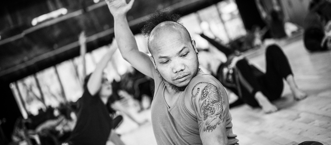 [Image Description: A black and white photo of Nadenh dancing in a studio. He sits on the floor and stretches one arm up behind him as the other remains on the floor. Nadenh is a disabled Cambodian man with light brown skin, he has a shaved head wth a black ponytail at the back, on his left upper arm he has a tattoo of a tiger. Other dancers stretch and reach whilst seated on the wooden floor in the background. 