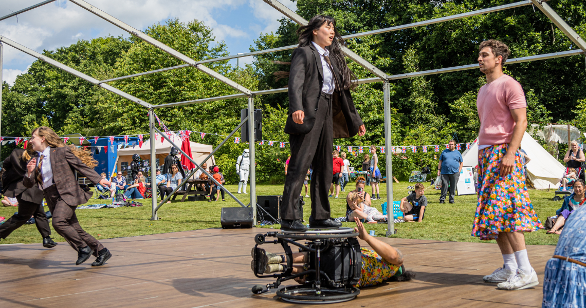 A performance of our outdoor work Frock at a Summer Fete event. Mid-show, Emily stands and spins on the upturned wheel of Nadenh’s wheelchair as he lays sideways on the ground. Other dancers in the six person ensemble run and stand in the background, audience sits and stands on grass around the flat wooden stage area. Emily wears a black suit, white shirt and tie. Christian stands to the right side, dressed in a light pink top, floral skirt and white trainers. 