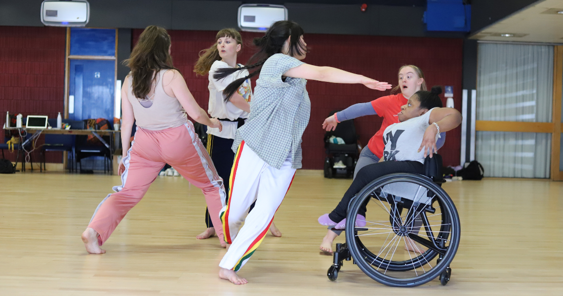 A moment in rehearsals of five dancers dancing closely weaving around one another in a studio. Emily is central in the image, titling off balance as she reaches a straight arm out towards wheelchair dancer Mo.