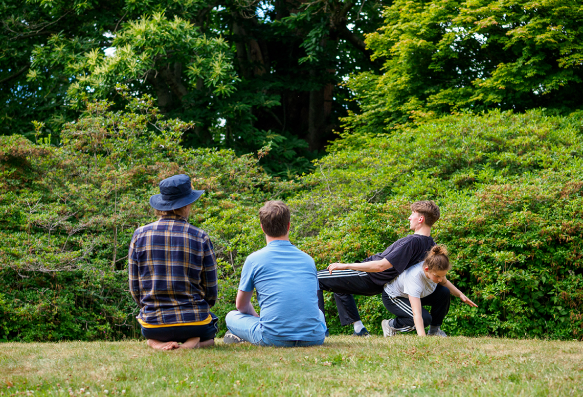 Tom and Chris sat side by side, backs to the camera and watching Abbie and Jonathan who rehearse a duet in front of them on a grassy bank. On the left, Tom, a white man, kneels and sits on his bare feet, on the right, Chris sits cross-legged. Abbie and Jonathan, two white standing dancers, are back to back leaning against one another but Abbie is crouched lower and Jonathan reclines back onto her a bit more upright. 
