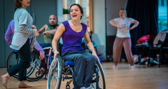 Laura in a studio, smiling widely as she travels across the space in a manual wheelchair. Laura is white with dark hair tied back from her face, she wears a purple t-shirt and grey trousers.