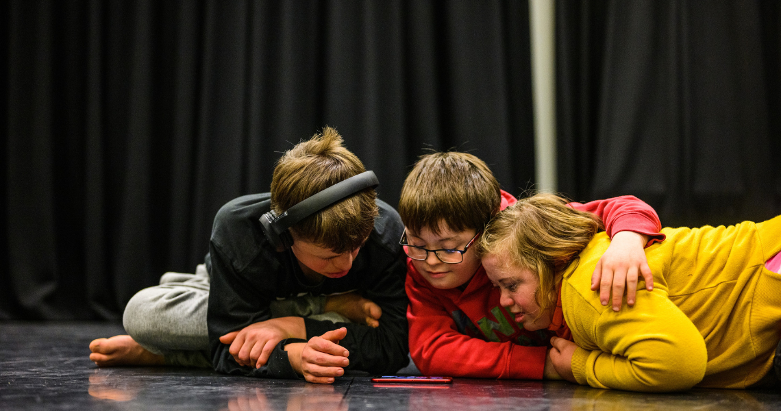 Three young white people with Down syndrome, huddled close on the floor they gather around a phone watching something together. On the left the young person sits cross legged and wears over-ear headphones, the central person wears glasses and they put their arm around their friend on the right. 