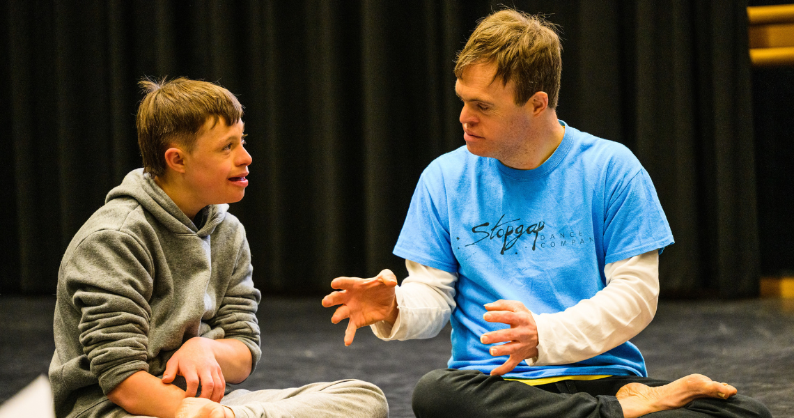 A young white person with Down Syndrome sits cross legged on the floor talking to Chris Pavia, a white man who also has Down syndrome. They smile at Chris as he talks to them, gesturing his hands.