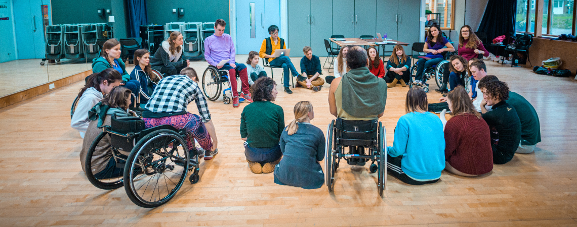 A large group of Stopgap staff and dancers gathered in a circle in a studio, people sit on the floor, on chairs and in wheelchairs as they listen and talk to one another. 