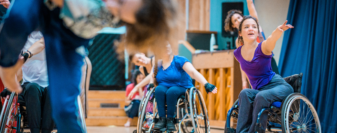Laura during a dance class, she reaches and points her left arm into the distance in front of her, other wheelchair dancers follow her in the background.  