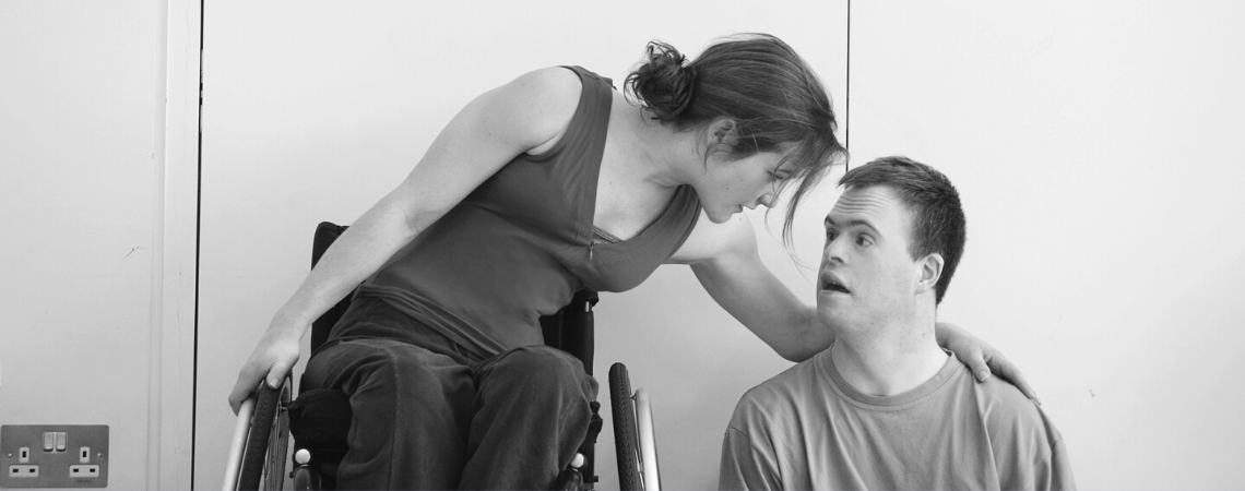 A black and white archive shot from the studio, Laura leans over to rest an arm round Chris' shoulder as they share a quiet conversation, she braces herself with her other hand on the right wheel of her chair. Chris is a white learning disabled man, Laura is a white disabled woman in a manual wheelchair. 