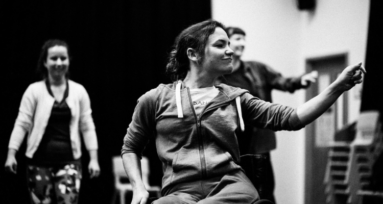 A black and white photo of Co-Artistic Director, Laura Jones, as she dances in the studio. One hand grips the wheel of her manual wheelchair, as she reaches to the other side to click her fingers. Her gaze follows her hand as she smiles confidently. In the blurred background more dancers watch.