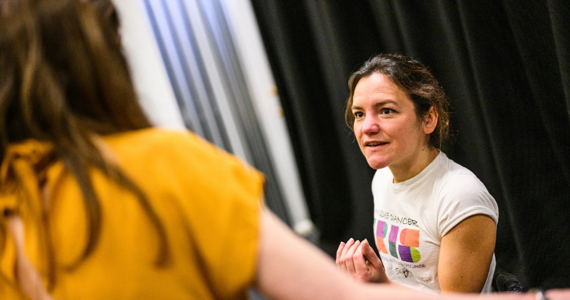 A close up of Co-Artistic Director, Laura Jones, teaching in a studio. Laura is talking to a figure in the foreground, facing away from the camera. She has her dark hair tied back, and is wearing a white T-shirt with the IRIS logo on as she sits in her wheelchair.