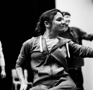 A black and white photo of Co-Artistic Director, Laura Jones, as she dances in the studio. One hand grips the wheel of her manual wheelchair, as she reaches to the other side to click her fingers. Her gaze follows her hand as she smiles confidently. In the blurred background more dancers watch.