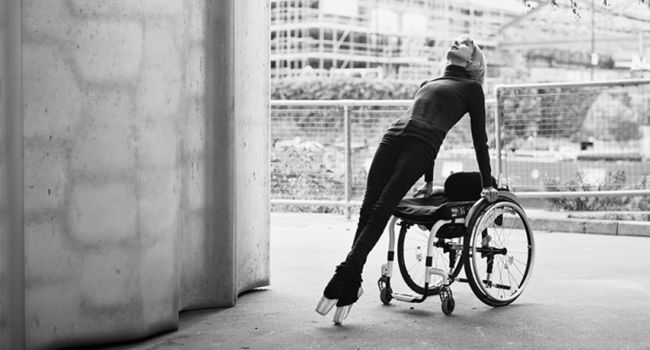A black and white image of Disabled dance artist Suzie Birchwood. Suzie stands and leans back onto her manual wheelchair.