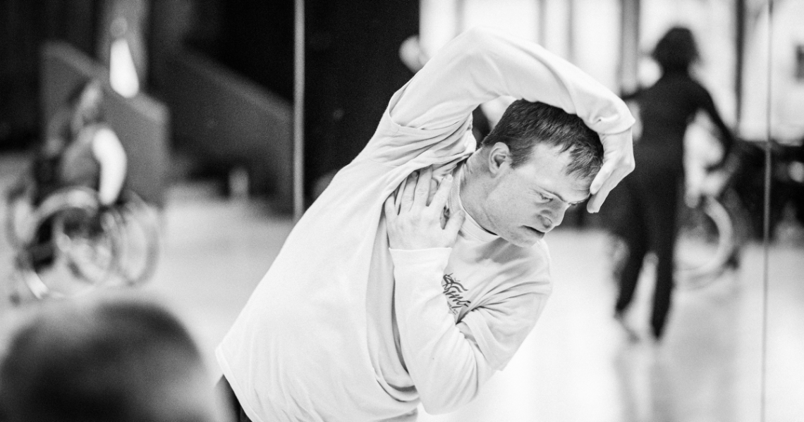 A black and white photo of Resident Choreographer, Chris Pavia. Chris is a white male dancer with Down syndrome. He curves his body to the side, one arm reaching over his head to rest on his forehead as he gazes serenely at the floor, and his other arm is folded up across his torso, hand resting below his neck