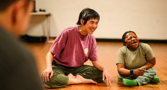 An east-asian woman and a black short-statured woman sit side by side and laugh together.