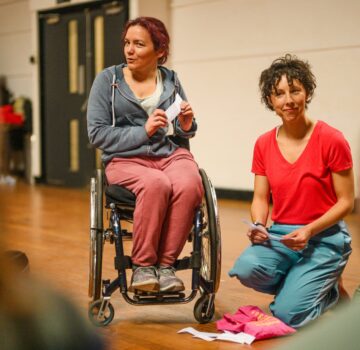 Two white women talking to a group, on the left the woman is a wheelchair user.