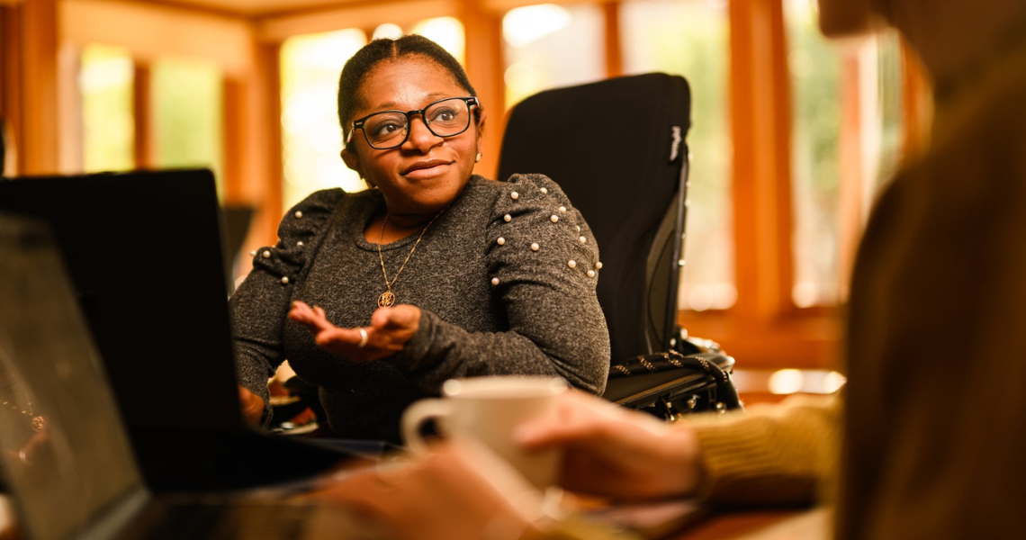 A black woman in a power wheelchair sits at a table and talks to someone out of frame.