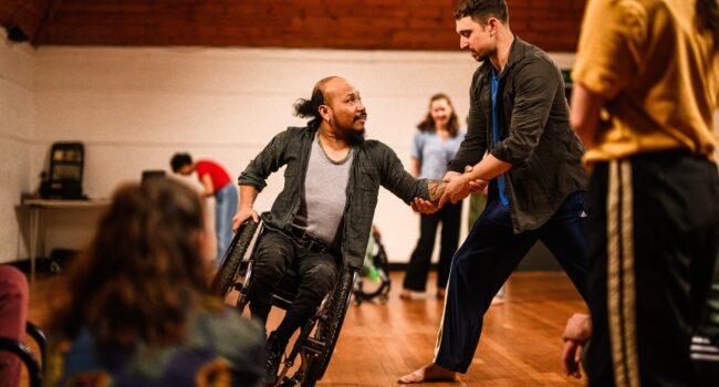 A wheelchair dancer tips to balance on their left wheel, and they place their hand into the support of a standing dancer.
