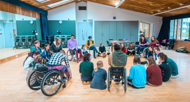 A large group of disabled and non-disabled people gathered in a circle in a large studio.