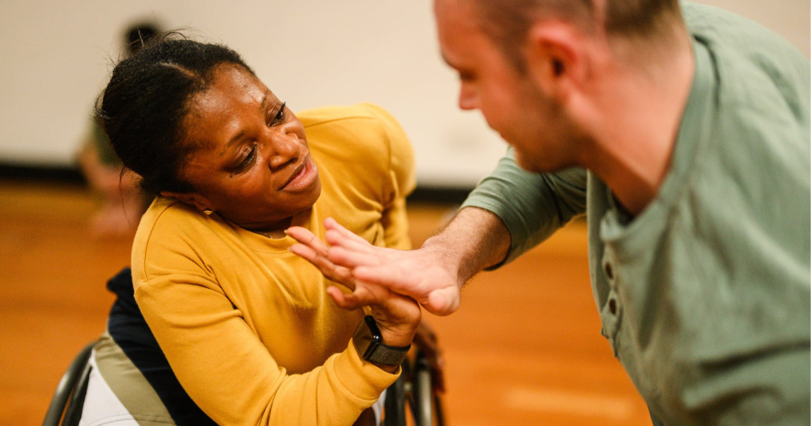A short-statured black woman in a manual wheelchair, presses their palm against another standing dancer's palm.