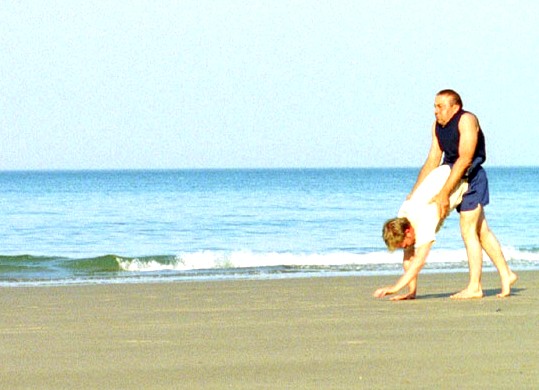A still from DV8's Cost of Living, Eddie Kay wanders along a beach on all fours, with Dave Toole on his back, giving the impression that Eddie's legs are Dave's.