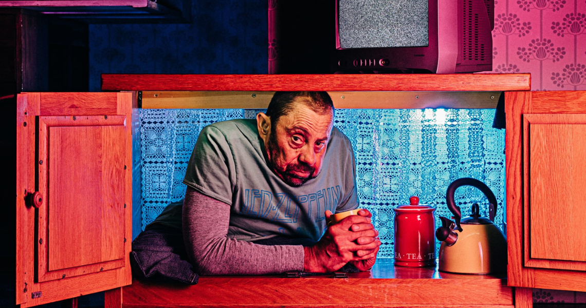 Dave on the set of The Enormous Room. Dave is white, has short dark hair and a beard, he is a disabled dance artist. He sits inside a wooden side cabinet with a lace curtain behind, clutching a mug in his remarkable hands. He rests on his forearms, a knowing look in his eyes inviting us in to his homely cupboard, his onstage world.