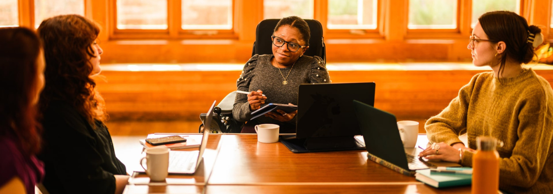 A black woman in a powered wheelchair sits at a table with an ipad and pen in her hands