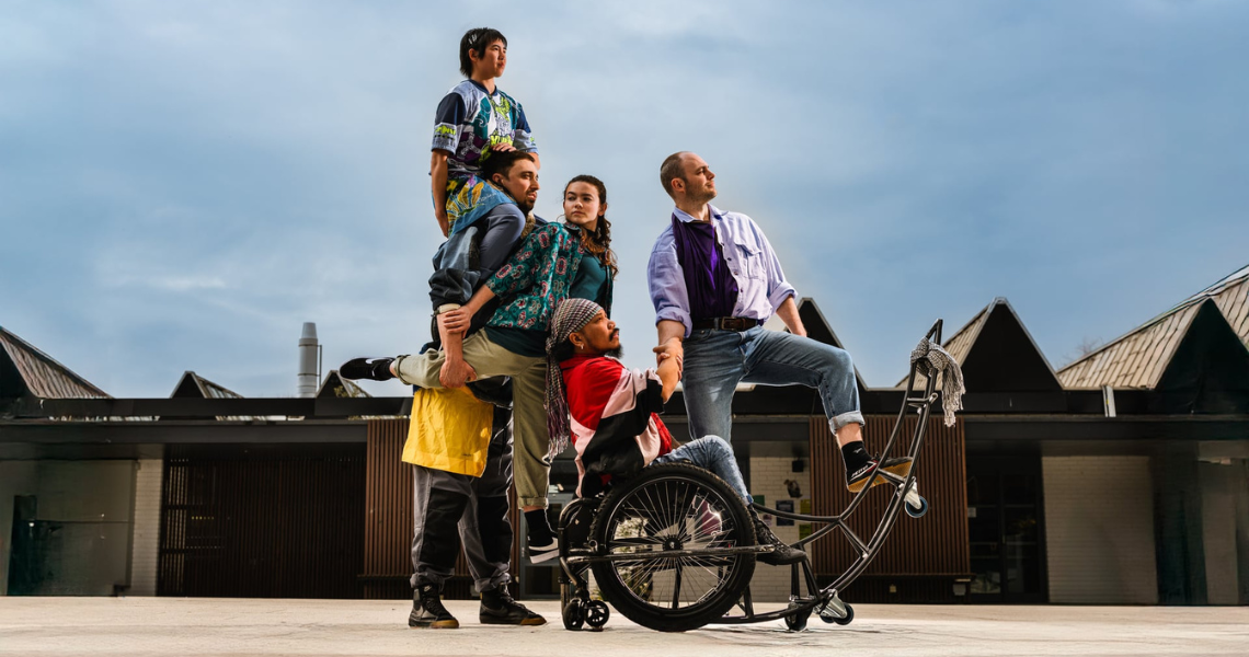 A group of five dancers outside. Gathered around Nadenh in his Ro-Tes, an adapted manual wheelchair with a long metal tail that curves upwards from his chair.