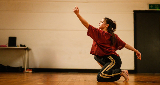 A white dancer in the studio, they crouch and reach a hand upwards.