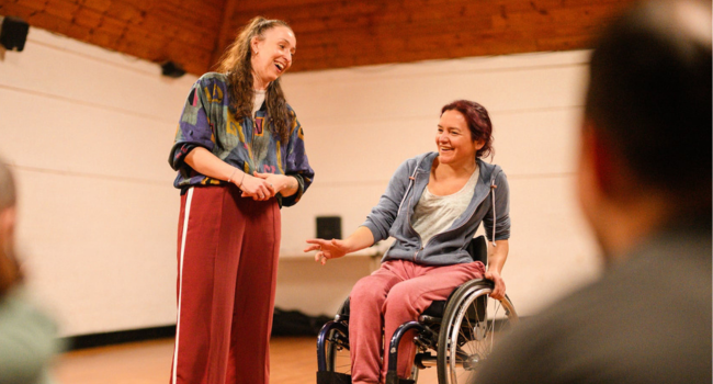 A non-disabled woman and a disabled woman speaking in front of a group.