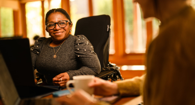 Monique, a Black disabled woman sits in her powerchair at a desk.