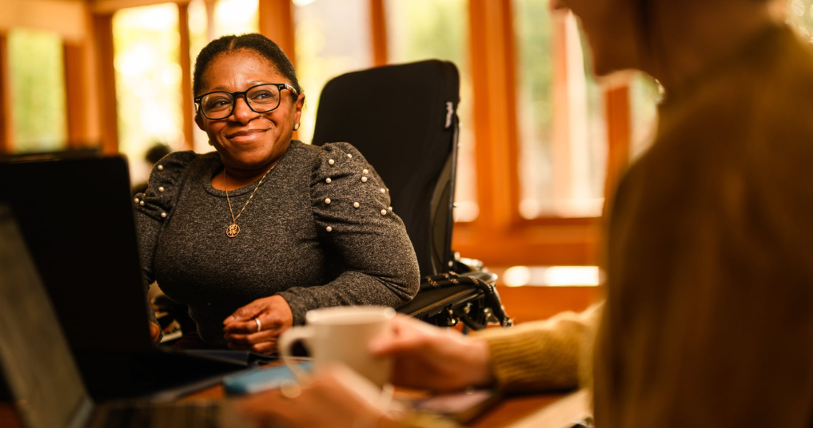 Monique, a Black disabled woman sits in her powerchair at a desk.